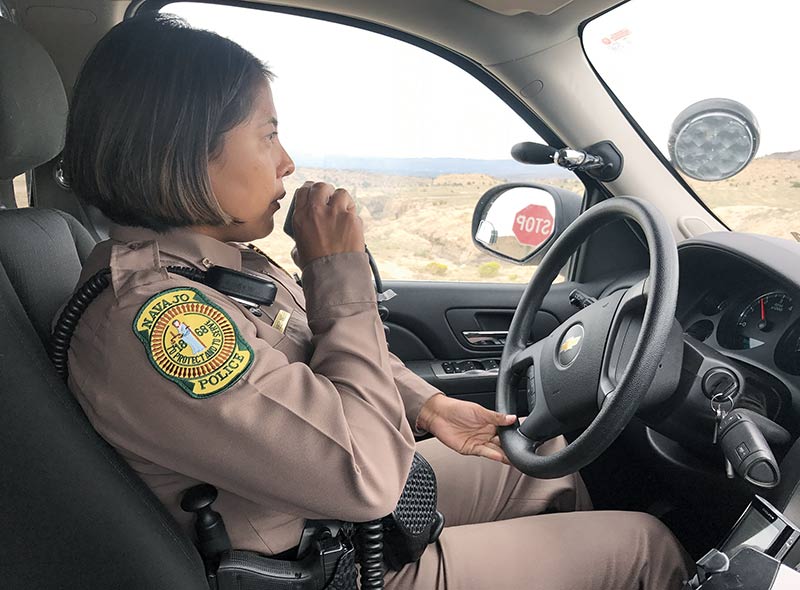 Police officer in car, driving, with radio microphone to mouth. High desert in background.
