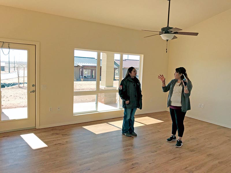 Two women in empty room with ceiling fan, large windows and wooden floor.